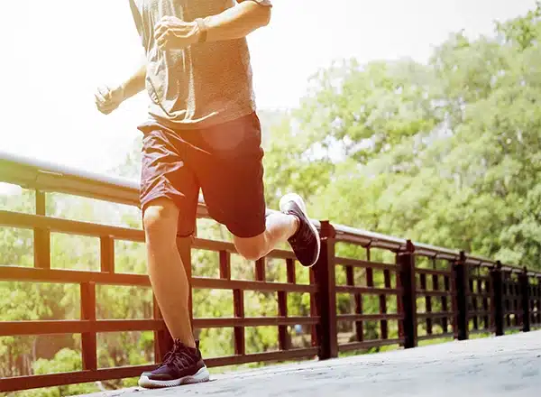 A mid-section shot of a person wearing a grey shirt, dark shorts, and black running shoes, running on a bridge railing on a bright, sunny day with green trees in the background.