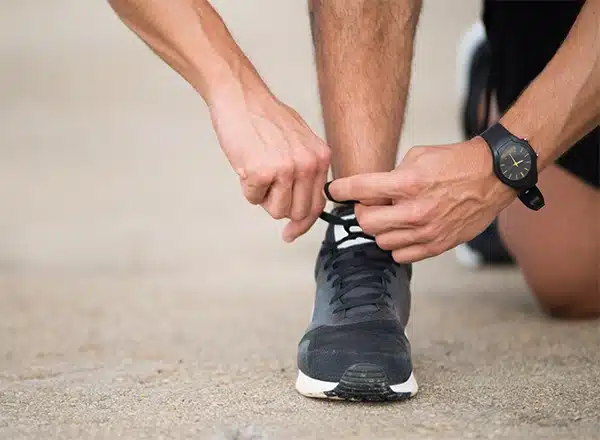 A close-up of a person's hands tying the black laces on a black running shoe on a concrete or paved surface. The person is wearing a black sports watch on their wrist.
