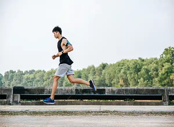 A full-body side profile of a male runner in a black tank top and grey shorts, running outdoors with an armband phone holder, on a paved surface next to a stone railing with green trees in the background.