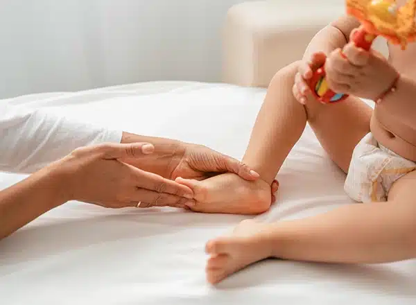 A close-up of a healthcare professional&rsquo;s hands gently palpating and examining the foot of an infant sitting on a white treatment table, while the baby holds a colorful toy.