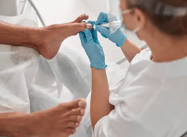 A pedicure technician wearing blue gloves and a face shield uses a professional electric nail file to treat a male client's toenails in a beauty salon.