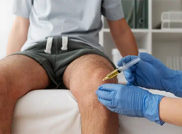 A close-up shot of a healthcare professional wearing blue gloves administering a syringe injection of yellow fluid into a patient's knee.