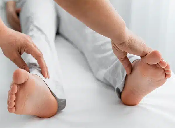 A close-up of a healthcare provider's hands manipulating the feet and ankles of a patient lying on a white examination table, likely performing a physical or osteopathic assessment.