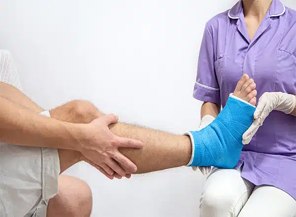 A medical professional in a purple uniform and white gloves holds a patient's foot, which is encased in a bright blue fiberglass cast or splint, during an examination.