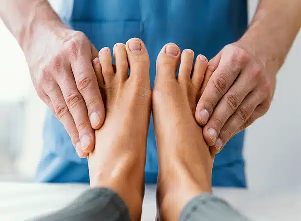 A close-up, first-person view of a patient's bare feet and toes being gently held and examined by a male osteopathic therapist wearing a blue uniform.