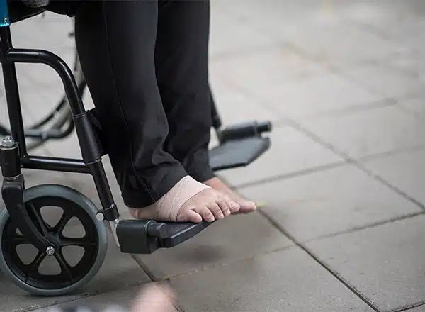 A close-up of an elderly person's feet, one wrapped in a light-colored bandage, resting on the footrest of a black wheelchair on a paved outdoor surface, illustrating foot or ankle pain.