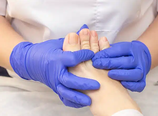 A close-up of a pedicurist or podiatrist wearing bright blue gloves, gently massaging or holding the bare foot and toes of a client or patient.