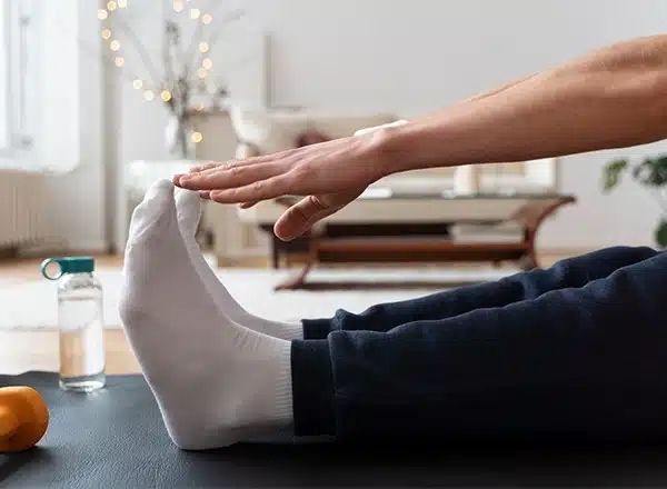 A close-up of a person sitting on a dark yoga mat in a brightly lit room, wearing white socks and dark pants, stretching forward to touch their toes. A small orange dumbbell and a glass water bottle are visible on the mat nearby.