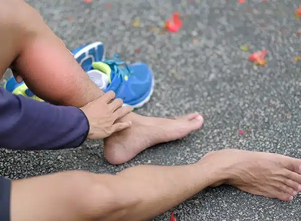 A close-up of a person sitting on an asphalt surface after a run, wearing a dark long-sleeve shirt and black shorts, holding their lower leg which is highlighted with a red glow indicating acute shin or calf pain. A bright blue and yellow running shoe rests nearby.