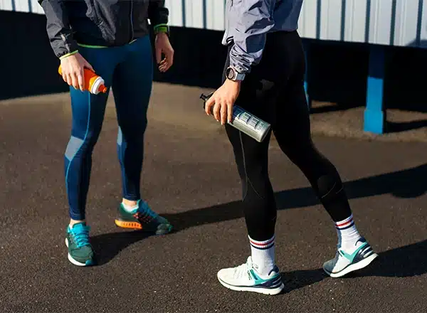 A low-angle, cropped image of two people wearing athletic gear and running shoes standing on asphalt, holding water bottles, after a workout, focusing on their legs and footwear.